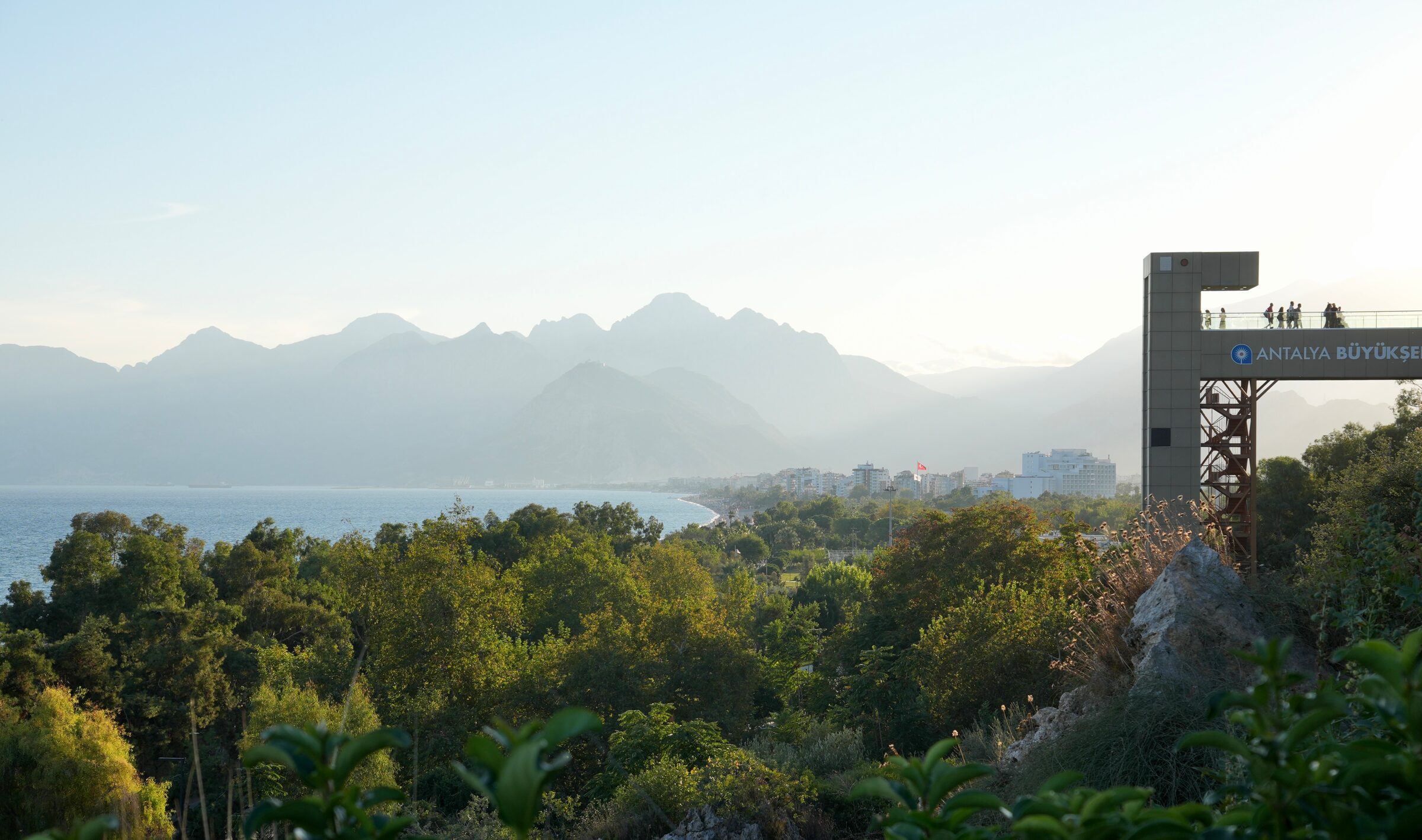 Blick über grüne Bäume auf die Küste von Antalya und die Berge im Abendlicht